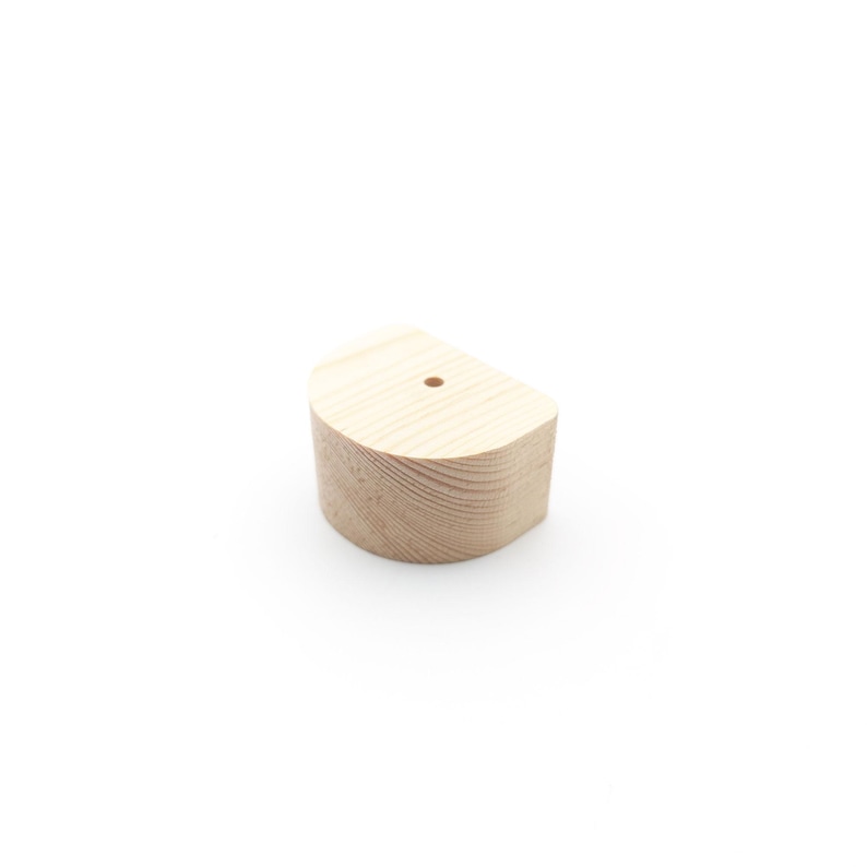 May include: A small, half-circle wooden furniture foot in natural wood tone. The foot has a flat base and a rounded top, with a small hole drilled in the centre. The wood grain is visible, and the piece is likely for DIY furniture projects.
