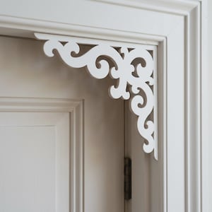 Può includere: Una staffa angolare in legno bianco caratterizzata da un intricato design a volute con motivi curvi e vorticosi. L'elemento decorativo è montato nell'angolo superiore di un telaio di porta, con una finitura liscia e intagli simmetrici.