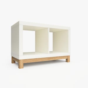 May include: White bookshelf with two open shelves and a wooden base. The bookshelf is made of wood and has a modern design.