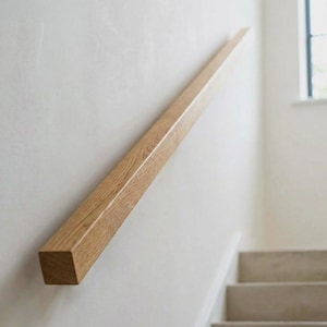 Könnte beinhalten: Ein helles Holzgeländer, das an einer weißen Wand befestigt ist. Das Geländer hat ein quadratisches Profil und eine natürliche Holzmaserung. Die Treppe ist im Hintergrund zu sehen. Das Geländer ist etwa 100 cm lang.