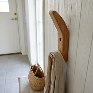 May include: A wooden coat rack fixed to a wall, featuring a curved design. A beige scarf is draped over the rack. A woven basket and a bench are visible in the background. The rack is made of light-coloured wood.