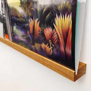 Może przedstawiać: Drewniany uchwyt ścienny do płyt winylowych. Uchwyt wykonany jest z naturalnego drewna i ma przezroczysty akrylowy wkład do ochrony okładki płyty. Okładka płyty przedstawia kolorową scenę dżungli w żywych odcieniach zieleni, żółci, czerwieni i fioletu.