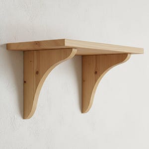 Op de afbeelding: Een lichtgekleurd houten plank met twee gebogen beugels. De plank is rechthoekig en lijkt van grenen te zijn. De beugels zijn ook van hout en hebben een decoratief, gebogen ontwerp. De plank is aan een witte muur bevestigd.