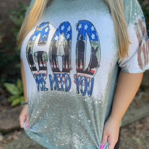 May include: Green t-shirt with a white bleached design. The design features the word "GOD" in large letters with an American flag pattern. Below the word "GOD" is the text "We Need You".