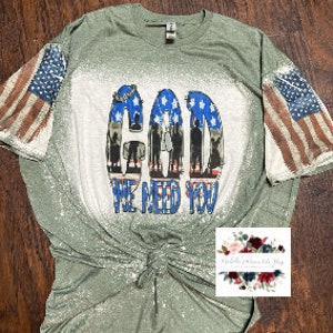 May include: Green t-shirt with a bleached design featuring the words "God We Need You" and an American flag on each sleeve.