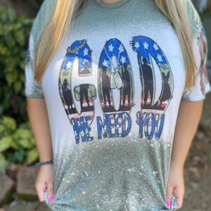 May include: Green and white t-shirt with a distressed look. The shirt features a graphic of the word "GOD" with a silhouette of soldiers in front of an American flag. The text "WE NEED YOU" is below the graphic.