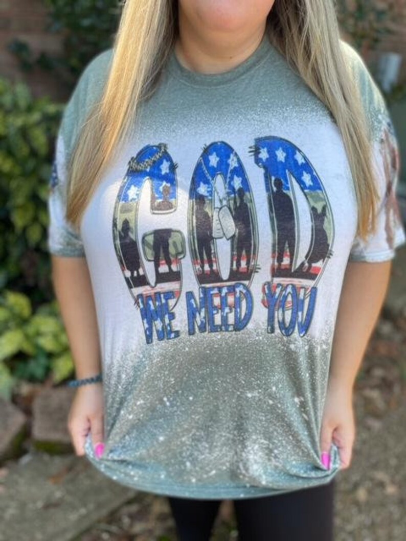 May include: Green and white t-shirt with a distressed look. The shirt features a graphic of the word "GOD" with a silhouette of soldiers in front of an American flag. The text "WE NEED YOU" is below the graphic.