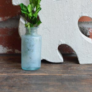 May include: A small, frosted blue glass vase holds a sprig of green leaves. The vase sits on a wooden surface, with a white, decorative wall hanging in the background. The wall hanging has a distressed, crackled finish.