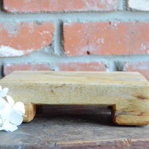 Könnte beinhalten: Ein kleiner, rechteckiger Holzaufsteller mit natürlichem Finish. Der Aufsteller hat ein schlichtes Design mit flacher Oberseite und kurzen Beinen. Links vom Aufsteller liegt ein Strauß weißer Blumen. Der Hintergrund ist eine Backsteinmauer.