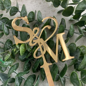 Può includere: Topper per torta in acrilico a specchio dorato con le lettere "S & M" in un font corsivo. Il topper è attaccato a un'asta di metallo dorato.