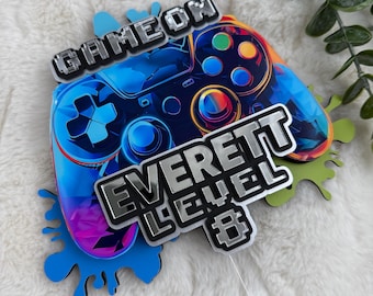 Könnte beinhalten: Ein farbenfroher Tortenaufsatz mit einem Videospiel-Controller-Design. Der Aufsatz trägt die Worte "GAME ON" und "EVERETT LEVEL 8" in silbernen Buchstaben. Der Controller ist blau mit bunten Akzenten.