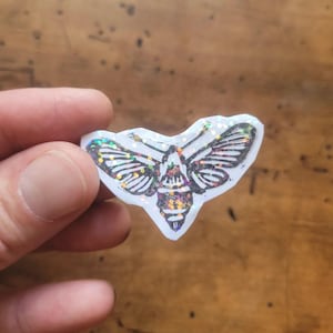 May include: A small sticker featuring a black and white line art illustration of a moth. The design is filled with iridescent, multi-colored glitter that reflects light, creating a sparkling effect across the wings and body of the insect.