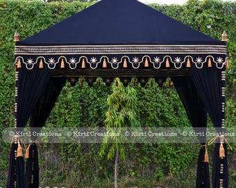 May include: A black canopy tent with gold trim and tassels. The tent is set up in a grassy area with trees in the background.