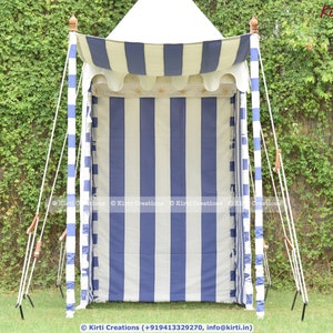 May include: A white and blue striped canopy tent with a white top and wooden poles. The tent is set up in a grassy area with green foliage in the background.