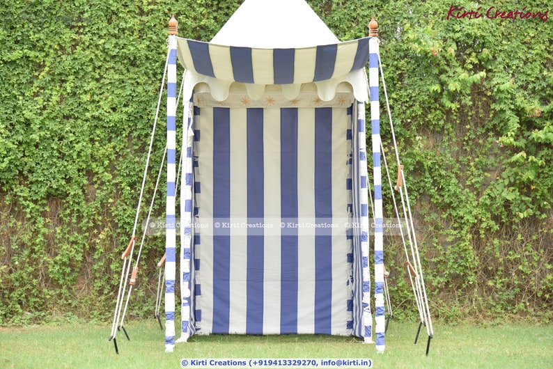 May include: A white and blue striped canopy tent with a white top and wooden poles. The tent is set up in a grassy area with green foliage in the background.