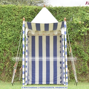 May include: A white and blue striped canopy tent with a white roof. The tent is set up in a grassy area with a green leafy wall behind it.