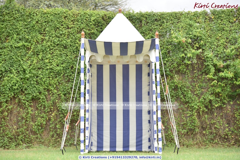 May include: A white and blue striped canopy tent with a white roof. The tent is set up in a grassy area with a green leafy wall behind it.