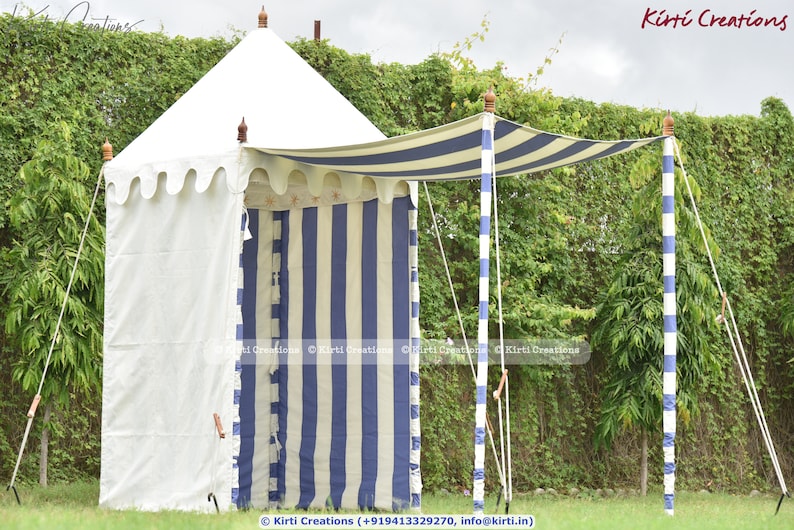 May include: A white and blue striped canopy tent with a white fabric wall. The tent has a wooden frame and is set up in a grassy area with green foliage in the background.