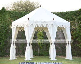 Puede incluir: Una carpa blanca con adornos decorativos y borlas. La carpa está instalada en un área de césped con vegetación en el fondo.