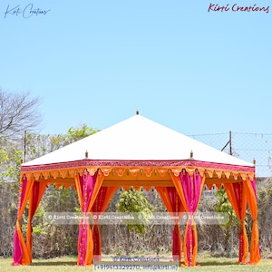 May include: A white, octagonal tent with orange and pink draping. The tent has a decorative trim and is set up on a grassy area. The text "Kirti Creations" is visible in the image.
