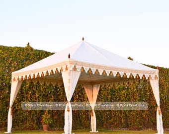 May include: A white canopy tent with gold trim and white curtains. The tent is set up in a grassy area with a green hedge in the background. The text "Kirti Creations" is visible in the image.