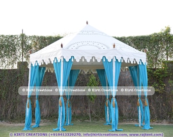 Puede incluir: Una carpa blanca con cortinas azules y borlas doradas. La carpa está decorada con encaje blanco y tiene un borde festoneado. La carpa está instalada en un área de césped con arbustos verdes en el fondo.