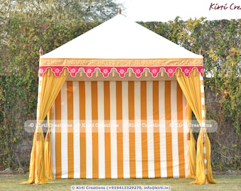 May include: A white and yellow striped tent with a pink and gold trim. The tent has a white canvas roof and yellow fabric curtains. The tent is set up in a grassy area with trees in the background.