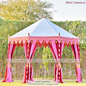 May include: A white and pink outdoor tent with decorative trim and draping curtains. The tent has a peaked roof and is set up on a grassy area. The text "Kirti Creations" is visible on the tent.