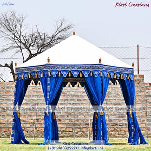 May include: A large outdoor pavilion tent featuring a white peaked roof and vibrant blue fabric curtains. The canopy edge is decorated with intricate gold and blue patterns, scalloped trim, and hanging gold tassels. The structure is supported by poles.