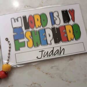 May include: A white card with the text "The Lord is my Shepherd" in colorful letters. The card has a hole punched in the top and a silver chain attached. The card also has a blank space for a name, which is filled in with the name "Judah".