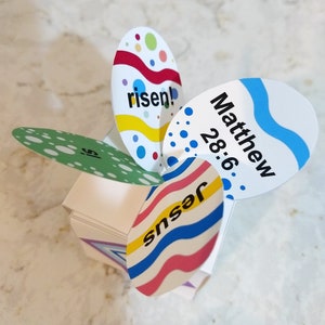 May include: A white paper Easter decoration with colorful egg-shaped cutouts. The cutouts feature the words "risen!" and "Jesus", and the text "Matthew 28:6". The decoration has a multi-layered design with a central base.