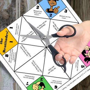 May include: A white paper craft with colorful geometric shapes and text from the Beatitudes, held by a person with black scissors. The paper has illustrations of people and phrases like "Blessed are the meek..." and "Blessed are the poor in spirit..."