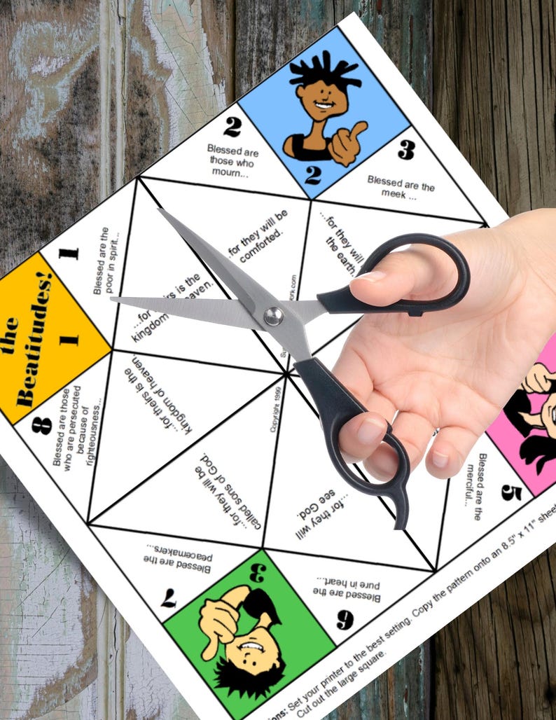 May include: A white paper craft with colorful geometric shapes and text from the Beatitudes, held by a person with black scissors. The paper has illustrations of people and phrases like "Blessed are the meek..." and "Blessed are the poor in spirit..."
