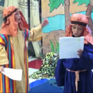 Puede incluir: Dos niños vestidos con disfraces, uno con una túnica amarilla y naranja con un patrón a rayas y el otro con una túnica azul, ambos con diademas naranjas. Están sosteniendo papeles y mirándose.