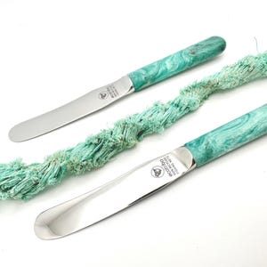 Peut inclure: Deux couteaux à tartiner en acier inoxydable avec des manches marbrés verts et blancs. Les lames sont argentées et les manches sont un tourbillon de vert et de blanc. Le texte "ecotibo 100% recycled fishing nets" est visible sur la lame.