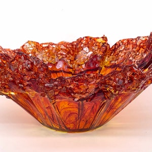 May include: A large, amber-colored glass bowl with a unique, textured, and irregular shape. The bowl has a wide, flared opening and a deep, rounded base. The glass is translucent and has a slightly iridescent quality.