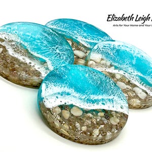 May include: Four oval resin coasters with a beach scene design. The coasters feature a turquoise blue wave design over a sandy beach base with seashells. The text "Elizabeth Leigh Arts" is visible in the upper right corner.