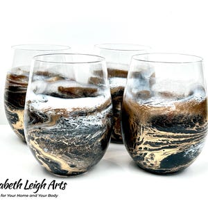 May include: Set of four wine glasses with a black, gold, and white marbled design. The glasses are clear and have a round shape.
