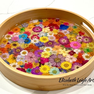 May include: A round bamboo tray filled with colorful dried flowers set in clear resin. The tray has two handles and the text "Elizabeth Leigh Arts" and "Arts for Your Home and Your Body" in black.