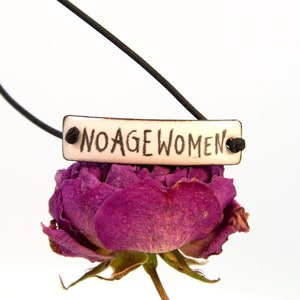 Könnte beinhalten: Ein weißer Keramik-Anhänger mit dem Text "NO AGE WOMEN" in schwarzen Buchstaben, der an einem schwarzen Band hängt und auf einer getrockneten violetten Rose liegt.