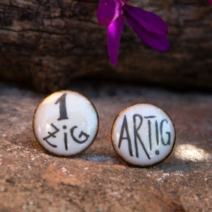 Op de afbeelding: Een paar ronde oorknopjes met een witte glanzende afwerking en donkere metalen randen. De linker oorknop bevat de tekst 1 ZIG in het zwart, terwijl de rechter oorknop de tekst ARTIG in het zwart toont, beide in een handgetekend lettertype.