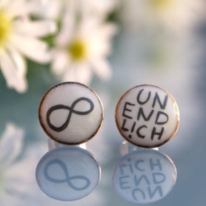 Op de afbeelding: Een paar witte ronde oorbellen met zwarte letters. De ene oorbel heeft een oneindigheidssymbool en de andere zegt "UNENDLICH".