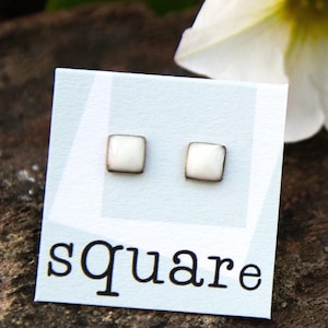 Könnte beinhalten: Ein Paar weiße quadratische Ohrringe mit silbernem Rand. Die Ohrringe werden auf einer weißen Karte mit dem Wort "square" in Schwarz gedruckt gezeigt.