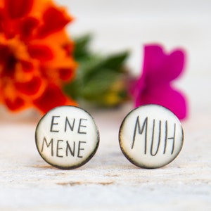 Könnte beinhalten: Ein Paar runde Ohrstecker mit cremefarbener Oberfläche und dunklen Rändern. Der linke Ohrstecker trägt den schwarzen Schriftzug ENE MENE, der rechte Ohrstecker den schwarzen Schriftzug MUH. Die Ohrstecker haben eine glatte, leicht gewölbte Oberfläche.