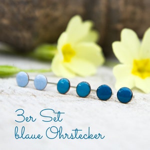 Op de afbeelding: Een set van zes blauwe oorstekers in verschillende tinten, gerangschikt in een rij. De oorbellen hebben een cirkelvormig ontwerp met een koperkleurige rand. De tekst "3er Set blaue Ohrstecker" staat in een turquoise script onderaan de afbeelding.