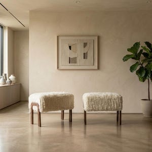 以下が含まれることがあります： 厚手のクリーム色のフェイクファー張りとダークブラウンの木製脚を備えた2つの長方形のオットマン。オットマンは、ベージュの壁、額装された抽象的なウォールアート、鉢植えのフィドルリーフフィグがある部屋に並べて配置されています。