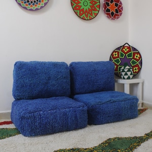 May include: Two blue, plush, square-shaped floor cushions with a white rug underneath. The cushions are arranged side-by-side, creating a small seating area.