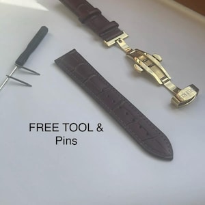 May include: Two brown leather watch straps with a gold-tone buckle. The buckle has the word "REIKO" engraved on it. The text "FREE TOOL & Pins" is written on a white surface.