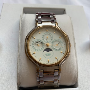 May include: A silver and gold Citizen Quartz watch with a white face and multiple subdials. The watch has a day, date, and moon phase display. The watch is in a white box.