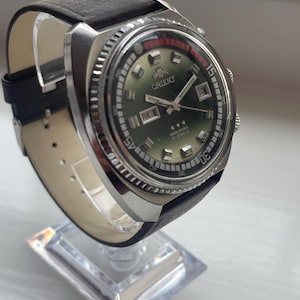 May include: A vintage Orient King Diver watch. It features a green dial, day and date display, and a black leather strap. The watch has a stainless steel case and a rotating bezel with red accents. The watch is displayed on a clear acrylic stand.